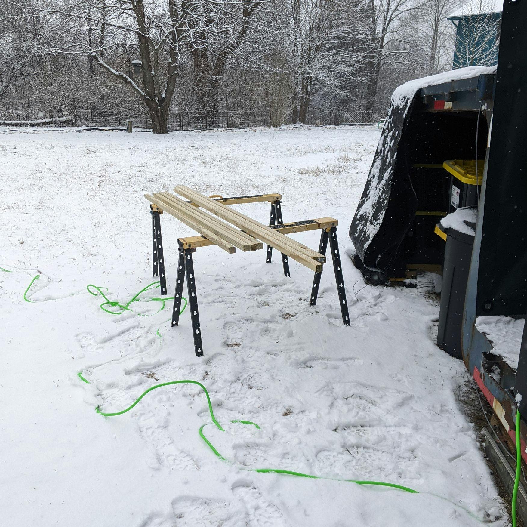 Sawhorses in the snow next to the tiny home with some 2x2s on them. The landlords snowy yard in the background